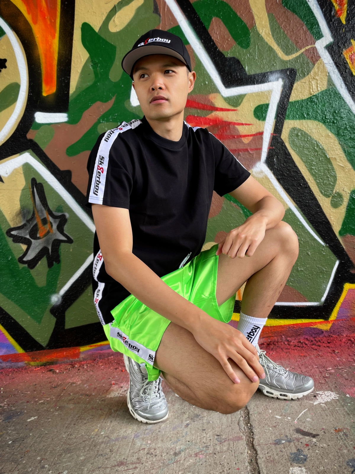 Model en short de sport Sk8erboy® vert fluo, T-shirt épais et chaussettes montantes devant un mur graffité