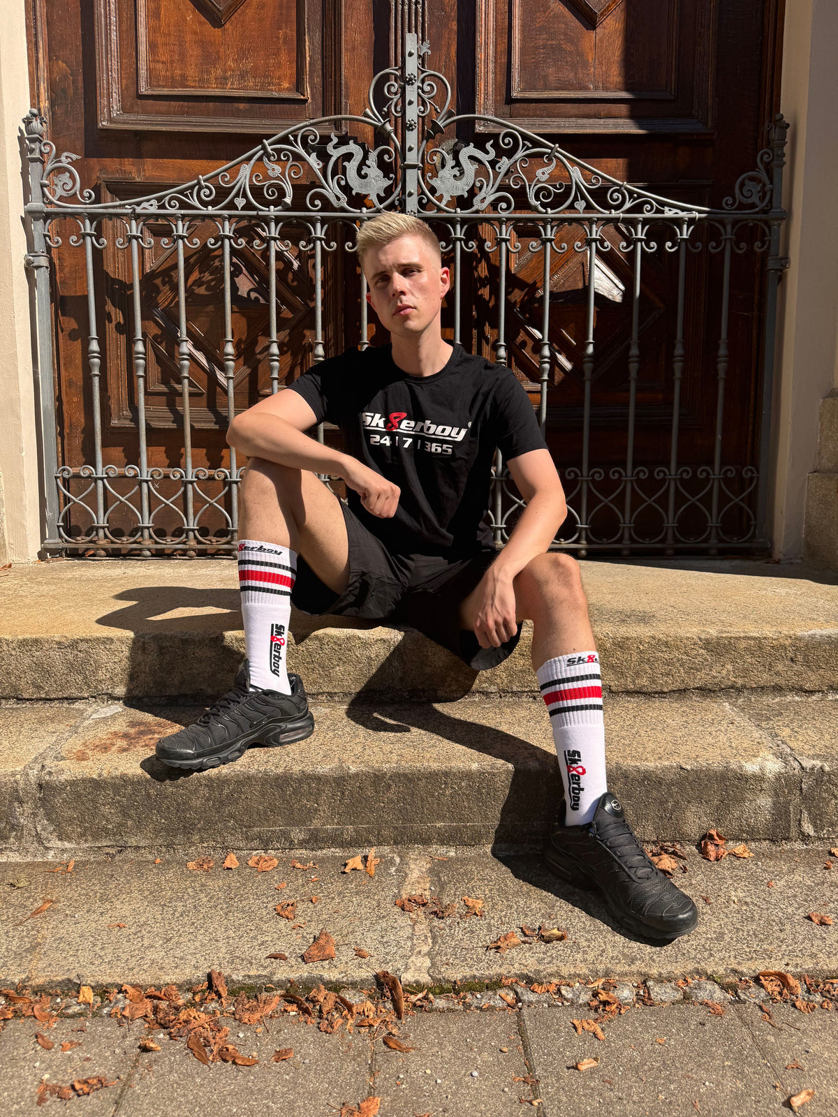 Joli twink gay portant un T-shirt Sk8erboy noir en coton, avec un imprimé Sk8erboy en lettres blanches et un 8 rouge, ainsi qu’un gros imprimé 24 7 365.