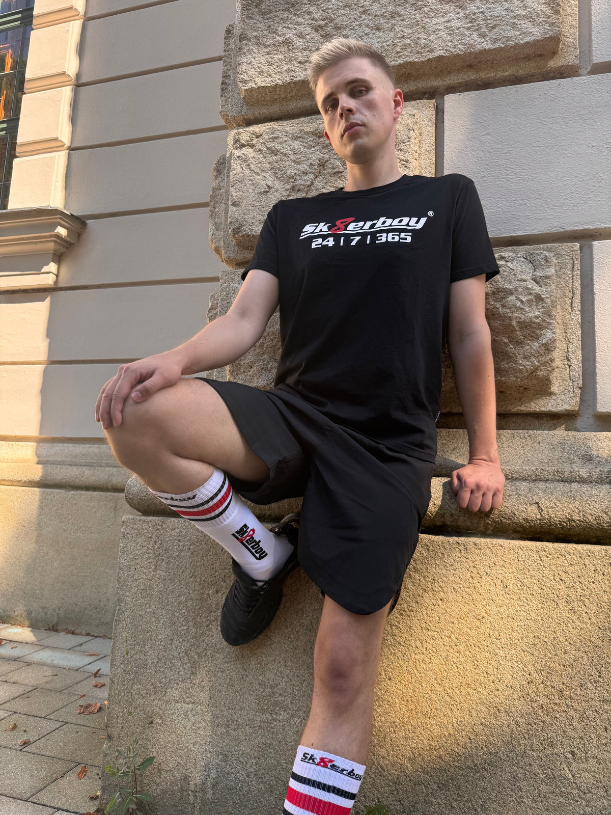 joli twink gay portant un t-shirt Sk8erboy noir en coton avec un imprimé Sk8erboy en lettres blanches et le chiffre 8 en rouge, avec un gros imprimé 24 7 365