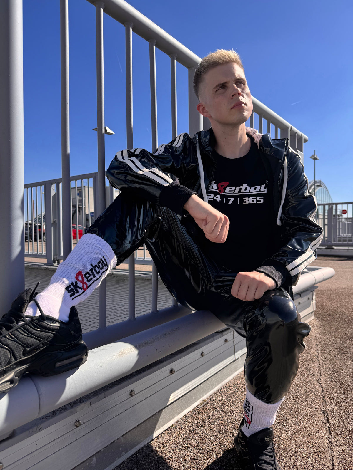 bel joli twink gay porte un t-shirt sk8erboy noir en coton avec un imprimé sk8erboy en lettres blanches et 8 en rouge, avec un gros motif 24 7 365 en tenue pvc