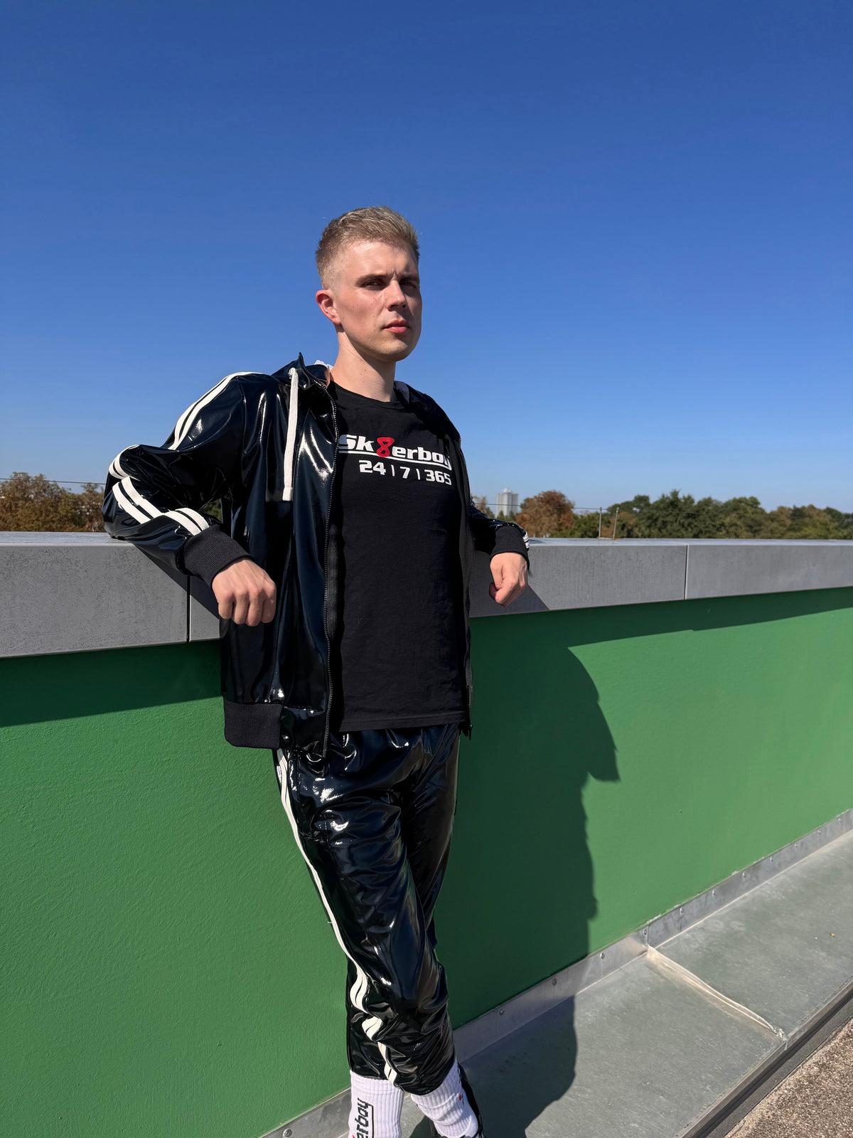 joli twink gay portant un t-shirt Sk8erboy noir en coton, avec un imprimé Sk8erboy en lettres blanches, un 8 rouge et un gros imprimé 24 7 365, ainsi qu’un vêtement en PVC