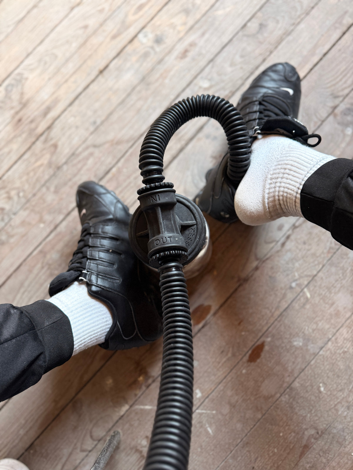 Deux pieds en shox en cuir noirs et chaussettes blanches, et sk8erboy avec un tuyau Vast Sniffer relié sur un sol en bois.