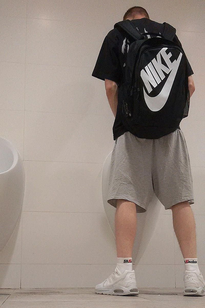 jeune homme gay en tenue de sport courte porte un sac à dos Nike et se tient devant un urinoir, avec des sneakers Nike et des chaussettes mi-hautes Sk8erboy blanches