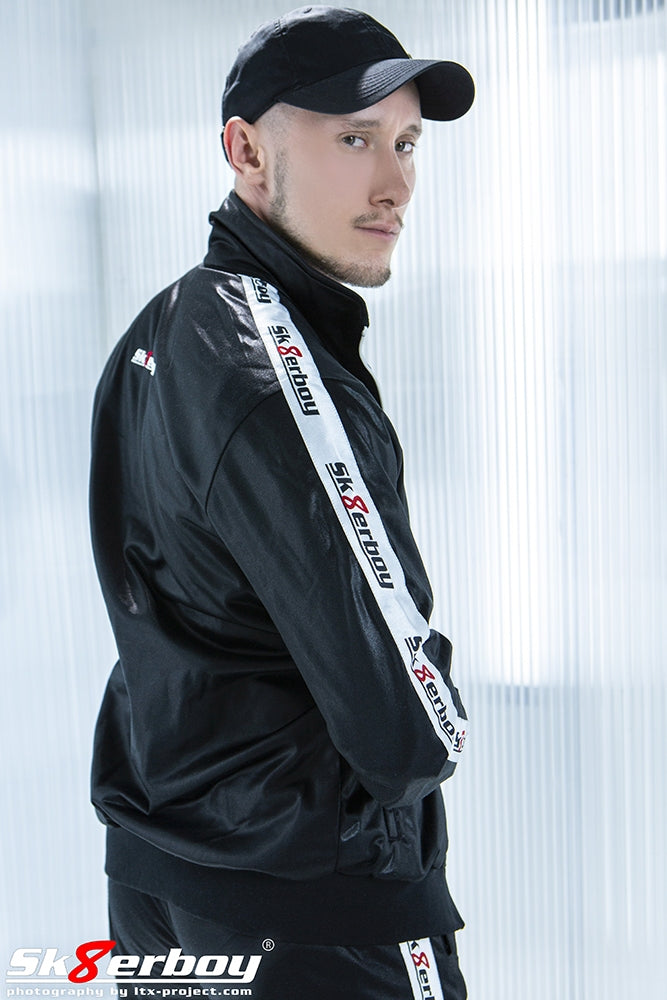 Jeune homme de côté, vu de dos, avec une veste shiny brillante de sk8erboy noire, une casquette et un pantalon shiny assorti.