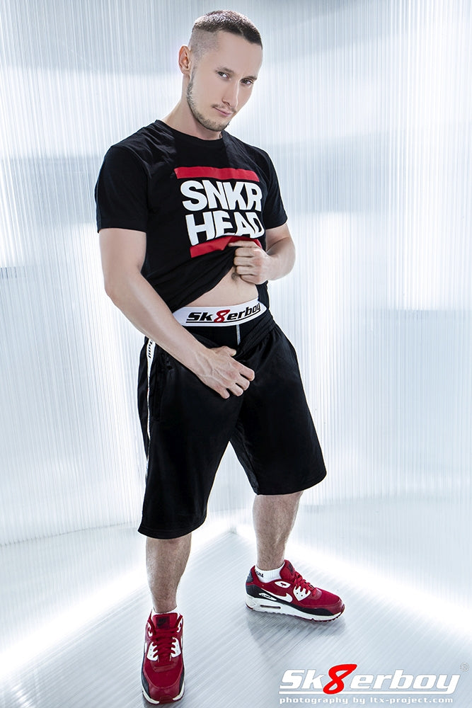 Jeune homme sportif portant un short brillant Sk8erboy et des sneakers Nike rouges, un T-shirt noir SNKR HEAD de Sk8erboy et un boxer assorti.
