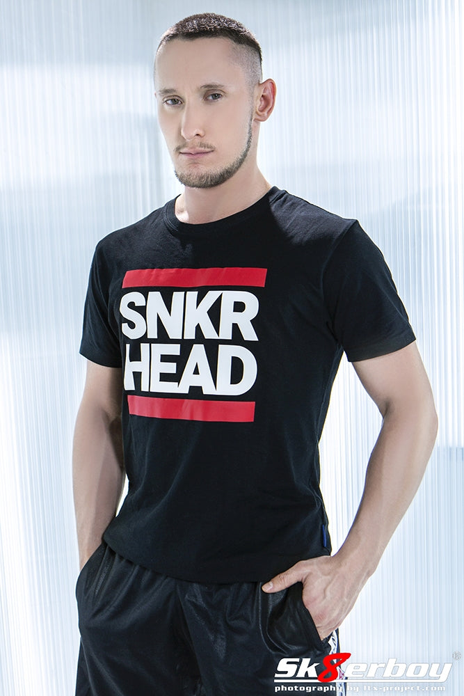 jeune gay portant un pantalon brillant de Sk8erboy et un t-shirt Snkr Head noir avec écriture blanche et bandes rouges