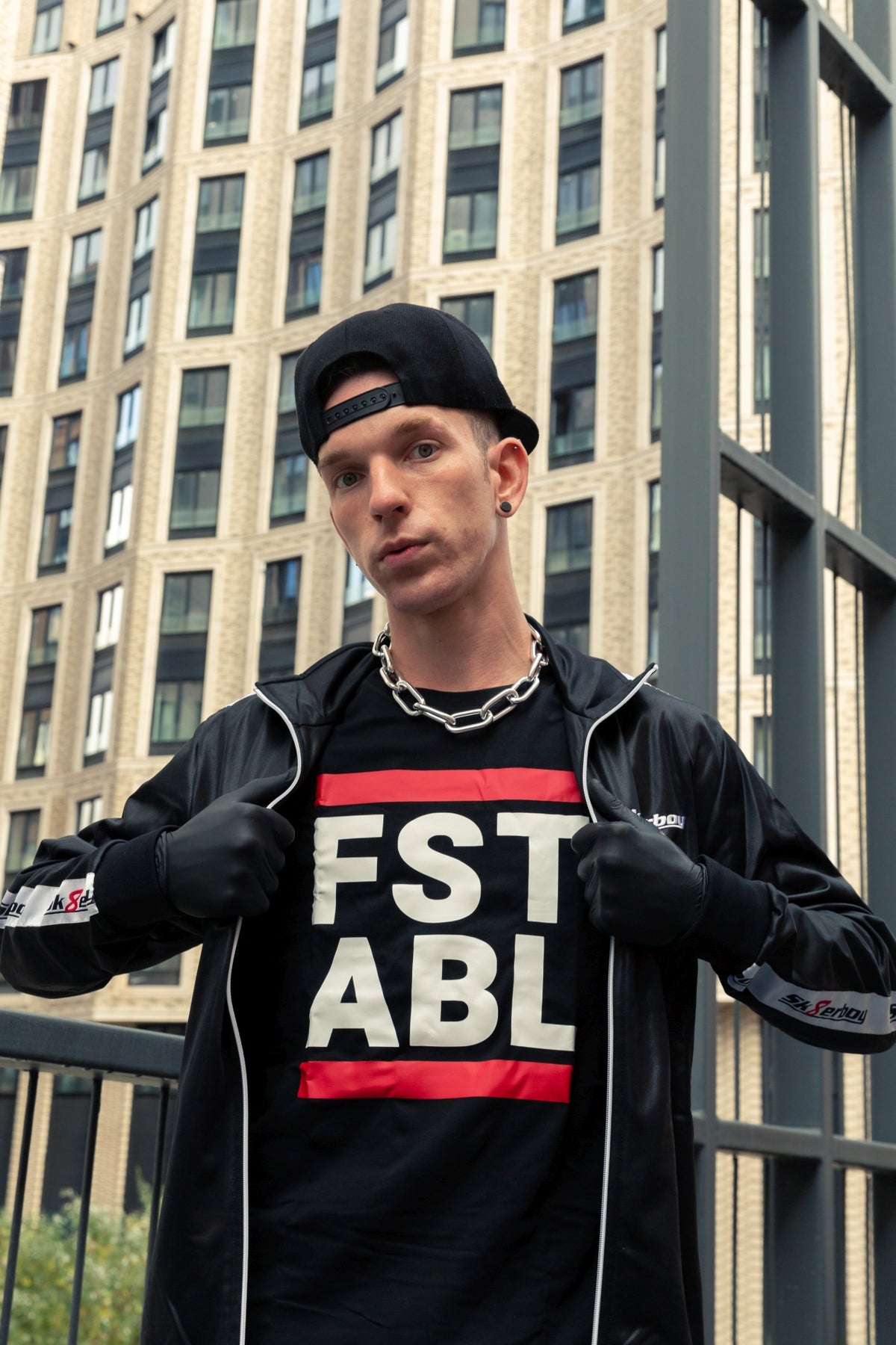 young gay man in the city wearing a black shiny tracksuit and a T-shirt with the FST ABL print, black gloves and a baseball cap matching the sk8erboy tracksuit