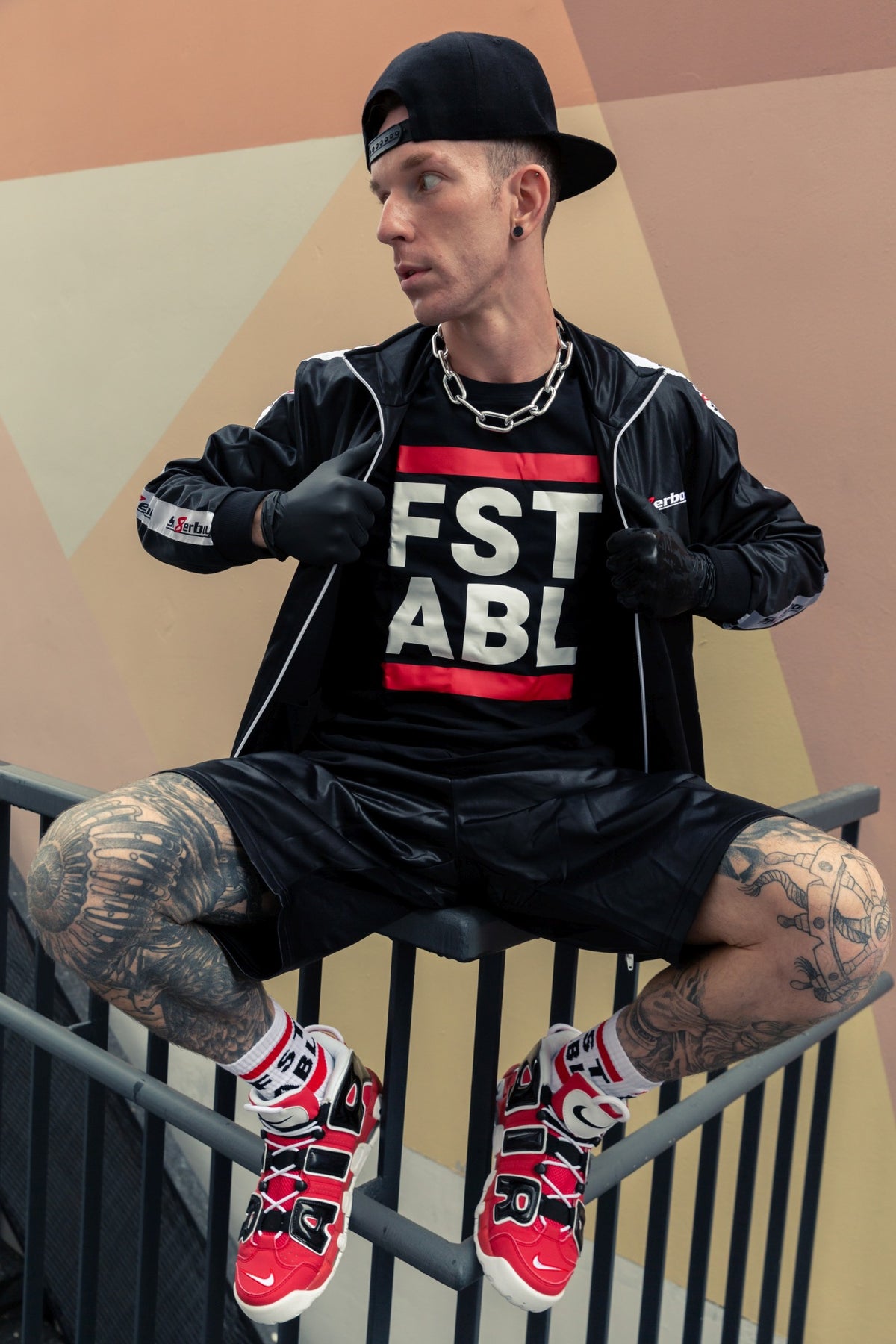 young man sitting on a railing wearing a shiny jacket from sk8erboy and matching shorts, with a black T-shirt with FST ABLE on the front and matching socks with red Nike Air sneakers