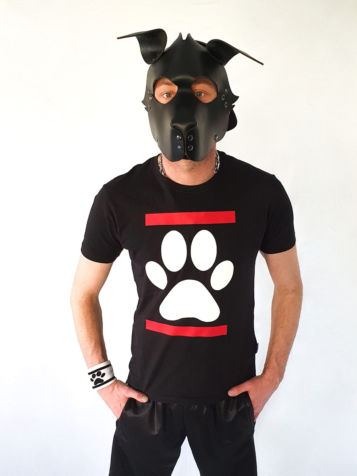 Jeune homme avec masque de chien en cuir et t-shirt noir Sk8erboy Dog Paw avec empreinte de patte blanche et pantalon de jogging brillant sur fond blanc