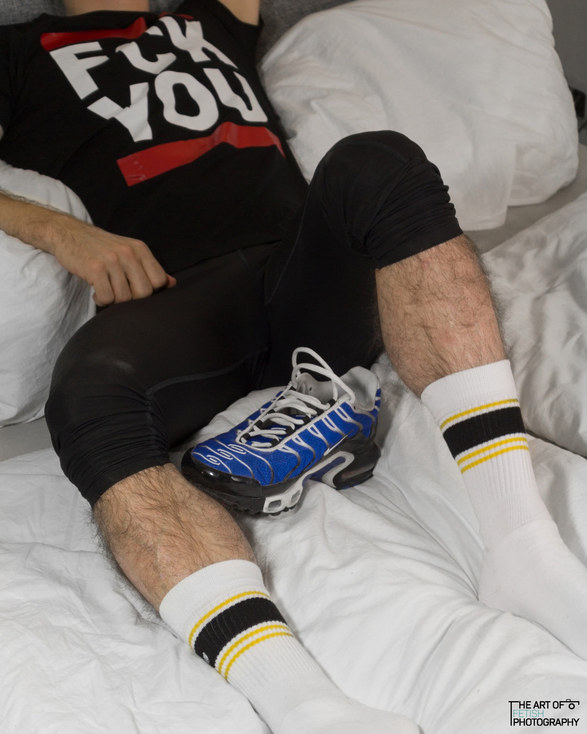 Jeune homme allongé dans le lit, en jogging et Nike TN, avec chaussettes Sk8erboy Deluxe jaunes, portant un T-shirt noir avec l’inscription « fck you » en blanc et bandes rouges.