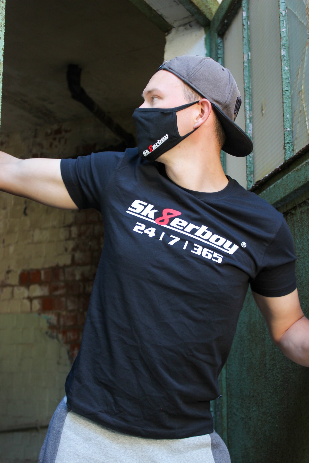 Un garçon porte un T-shirt sk8erboy noir en coton avec imprimé sk8erboy en blanc et un 8 rouge, «24/7/365» en gros. Il a aussi un masque sk8erboy, une casquette, un jogging gris, des Nike Air Max, devant une vieille usine abandonnée.