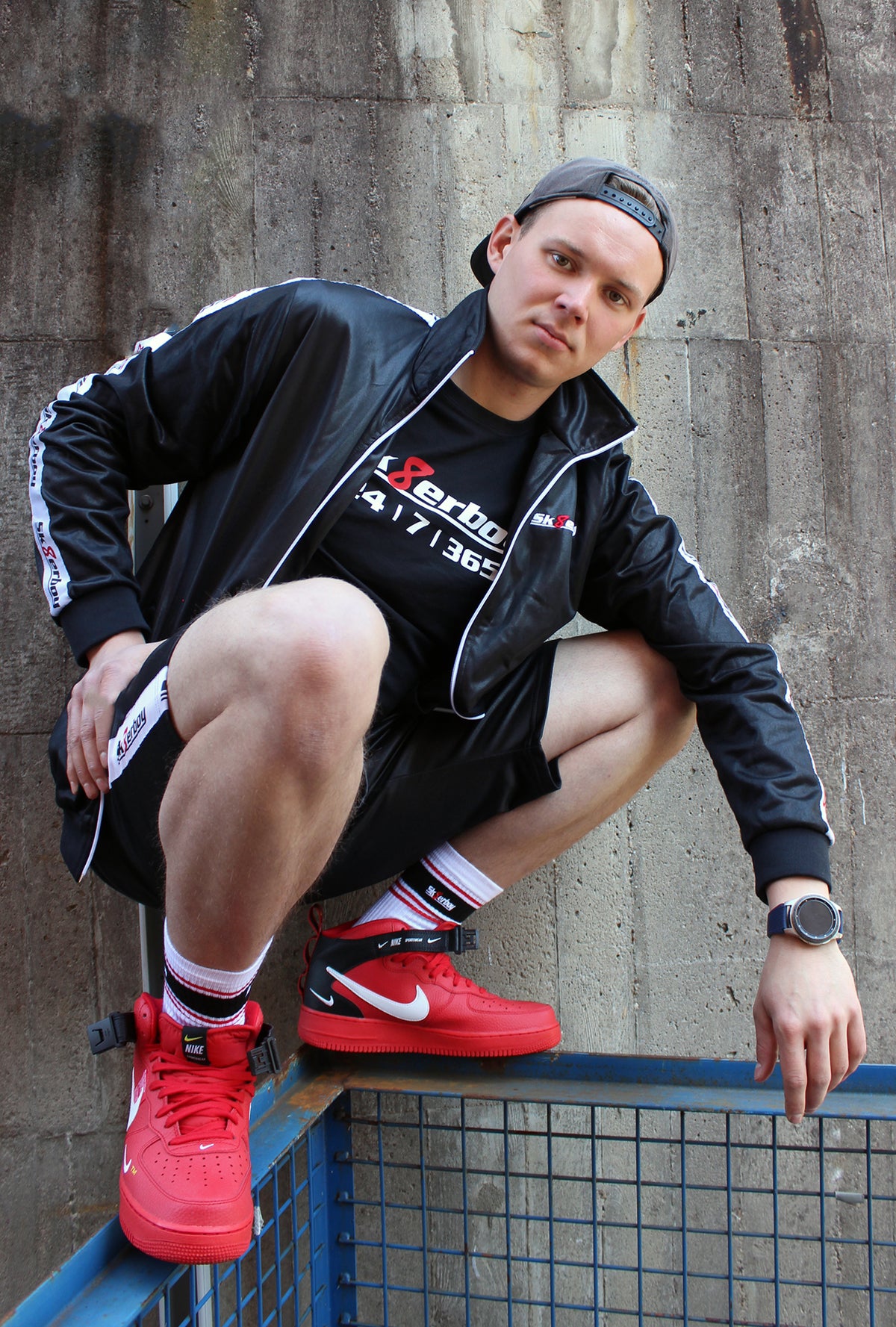 Un garçon porte un t-shirt sk8erboy noir en coton avec logo en lettres blanches, un 8 rouge et l’imprimé 24/7/365. Il porte aussi un masque sk8erboy, une casquette, un jogging gris Nike Air Max, devant une vieille usine abandonnée.