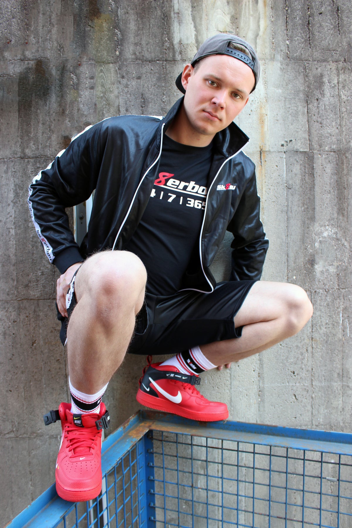 Un garçon porte un t-shirt Sk8erboy noir en coton, avec logo Sk8erboy en blanc, un 8 rouge et 24 7 365 en gros, masque Sk8erboy, casquette de baseball, jogging gris, Nike Air Max, devant une vieille usine abandonnée.