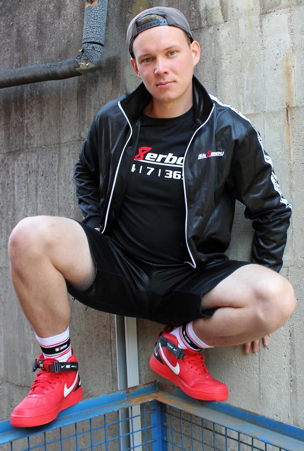 un garçon porte un T-shirt Sk8erboy noir en coton avec imprimé Sk8erboy, texte blanc et 8 rouge, 24/7/365 en gros. Il porte aussi un masque Sk8erboy, une casquette de baseball, un jogging gris, des Nike Air Max, devant une vieille usine abandonnée.
