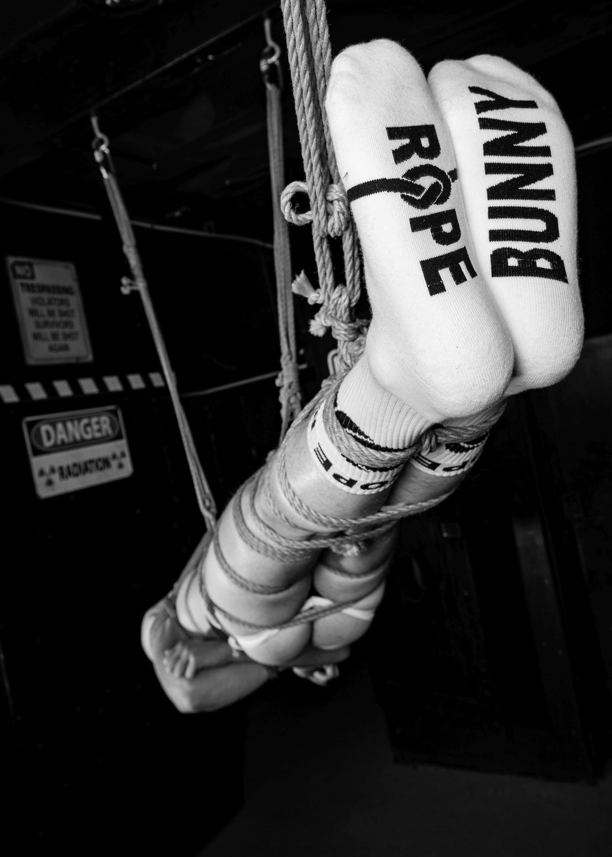 Bound young man hanging in ropes with sk8erboy rope bunny socks in white in front of a black wall in black and white