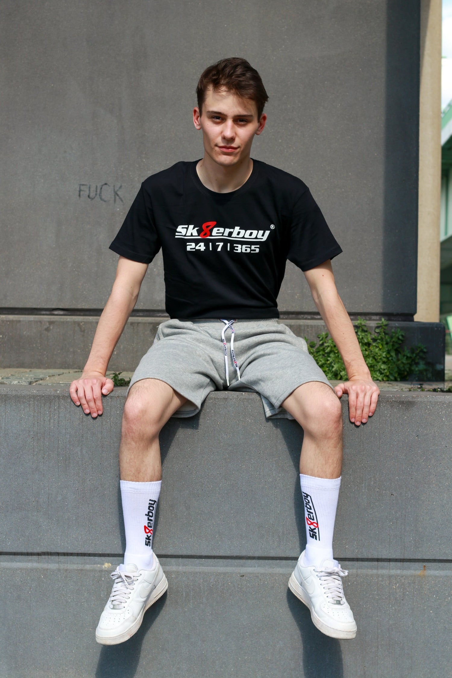 Joli twink gay porte un T-shirt Sk8erboy noir en coton, avec logo Sk8erboy en blanc, 8 rouge et gros imprimé 24/7/365, avec survêtement court gris Sk8erboy.