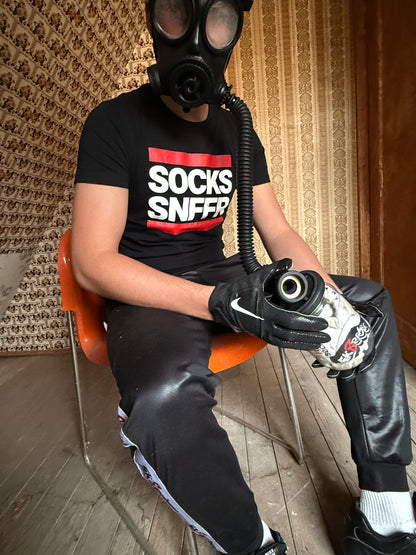 Person mit Gasmaske, schwarzem „Socks Snffr“-T-Shirt und Handschuhen sitzt auf einem orangefarbenen Stuhl und hält ein Sk8erboy Vast Sniffer Glas in der Hand