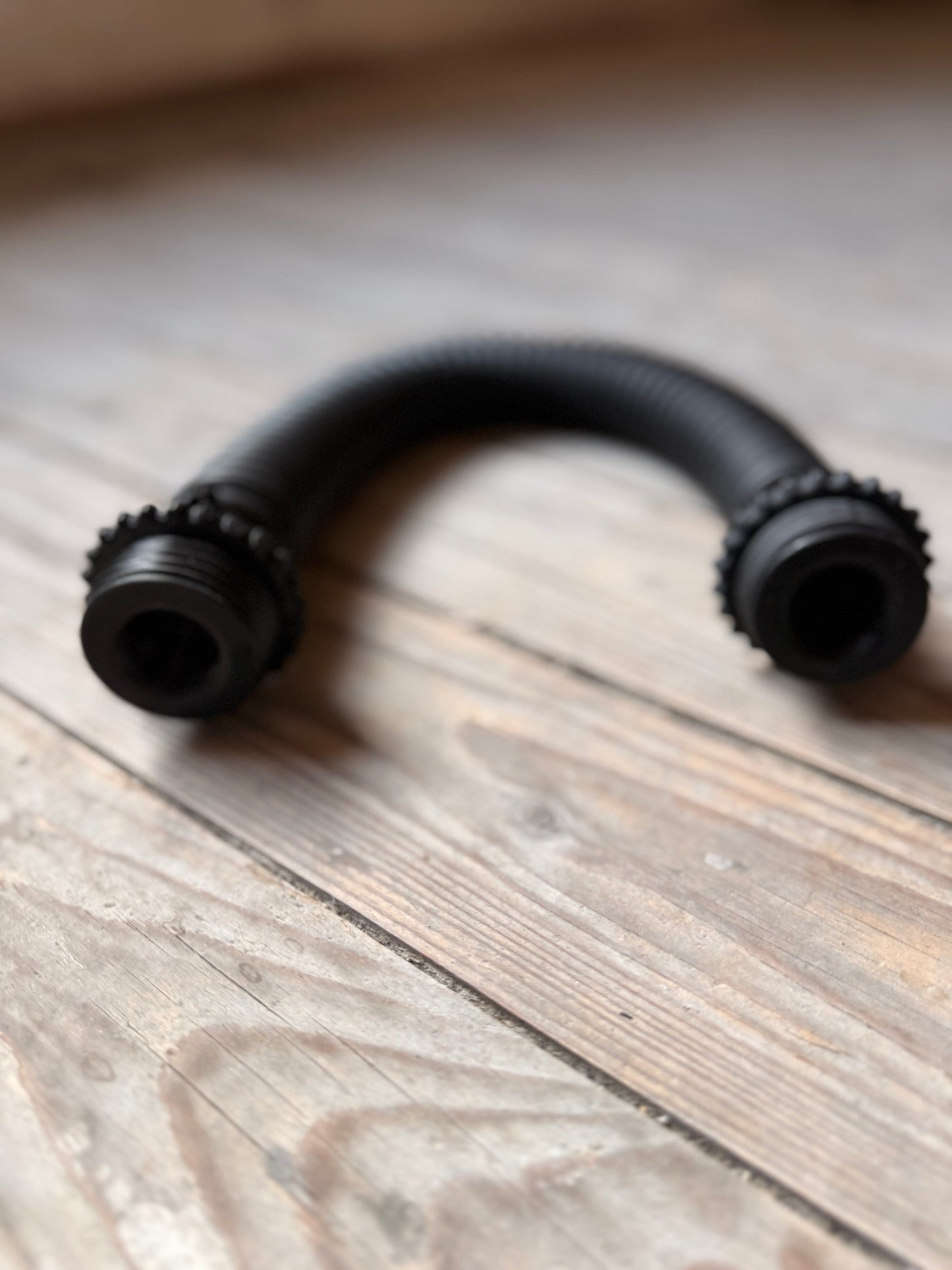 Schwarzer, flexibler Vast Sniffer Schlauch liegt gebogen auf einem Holzboden.