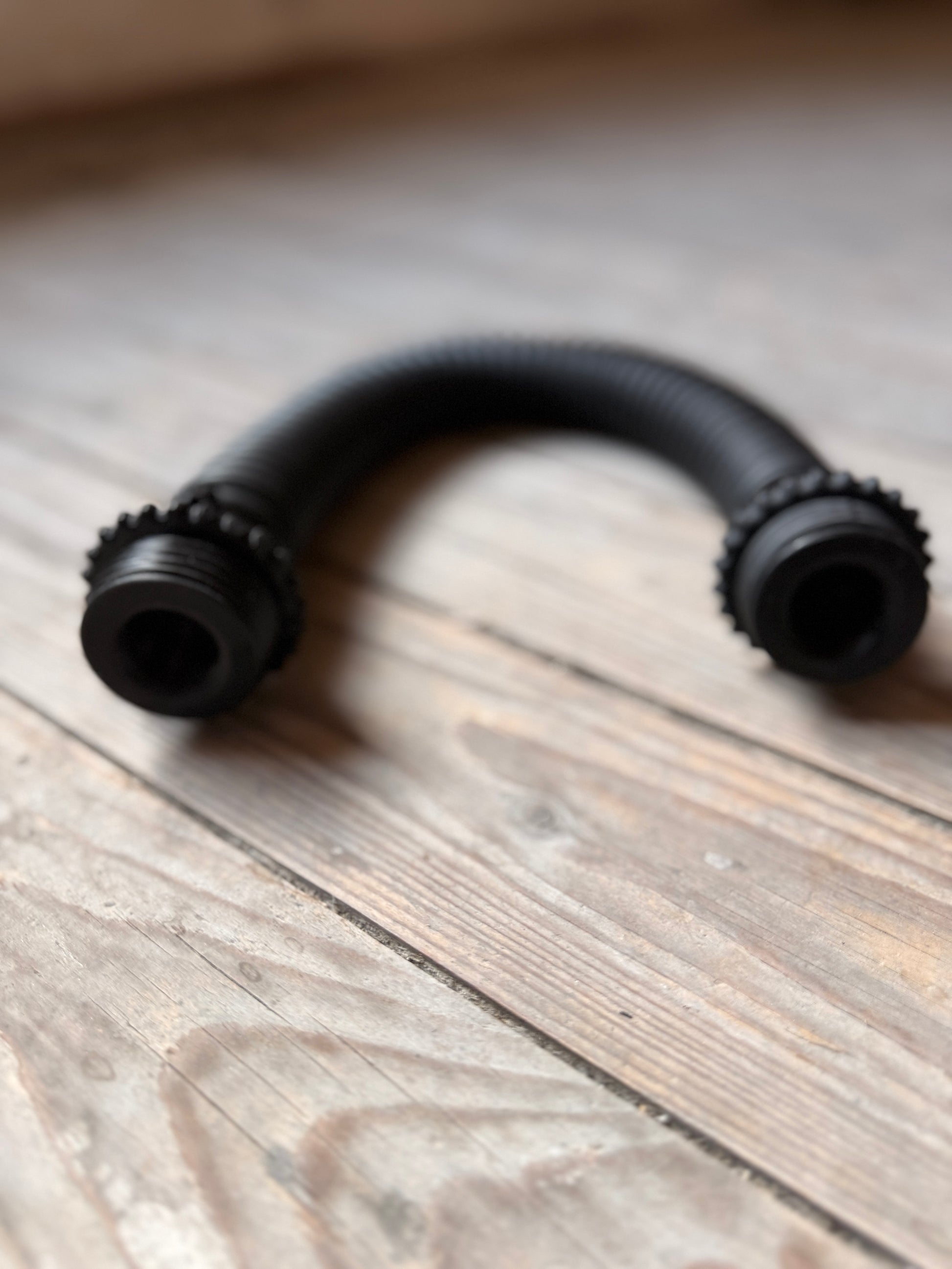 Schwarzer, flexibler Vast Sniffer Schlauch liegt gebogen auf einem Holzboden.